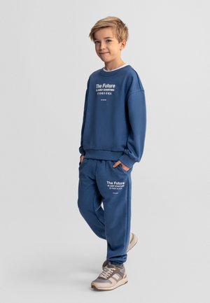 Jonge jongen staat met handen in zakken, draagt een blauwe sweatshirt en joggingbroek met de tekst "The Future is Just Starting", en sneakers, op een grijze achtergrond.