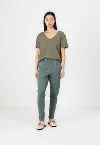 Olive green v-neck t-shirt, loose fit, paired with slim-fit, drawstring dark green trousers. Model wears beige flat shoes with decorative accent.