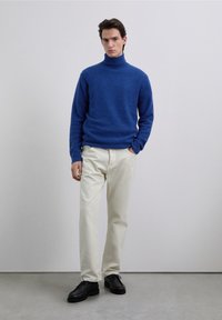 Maglione a collo alto blu, tessuto morbido, abbinato a pantaloni dritti color crema e scarpe nere stringate su sfondo neutro.