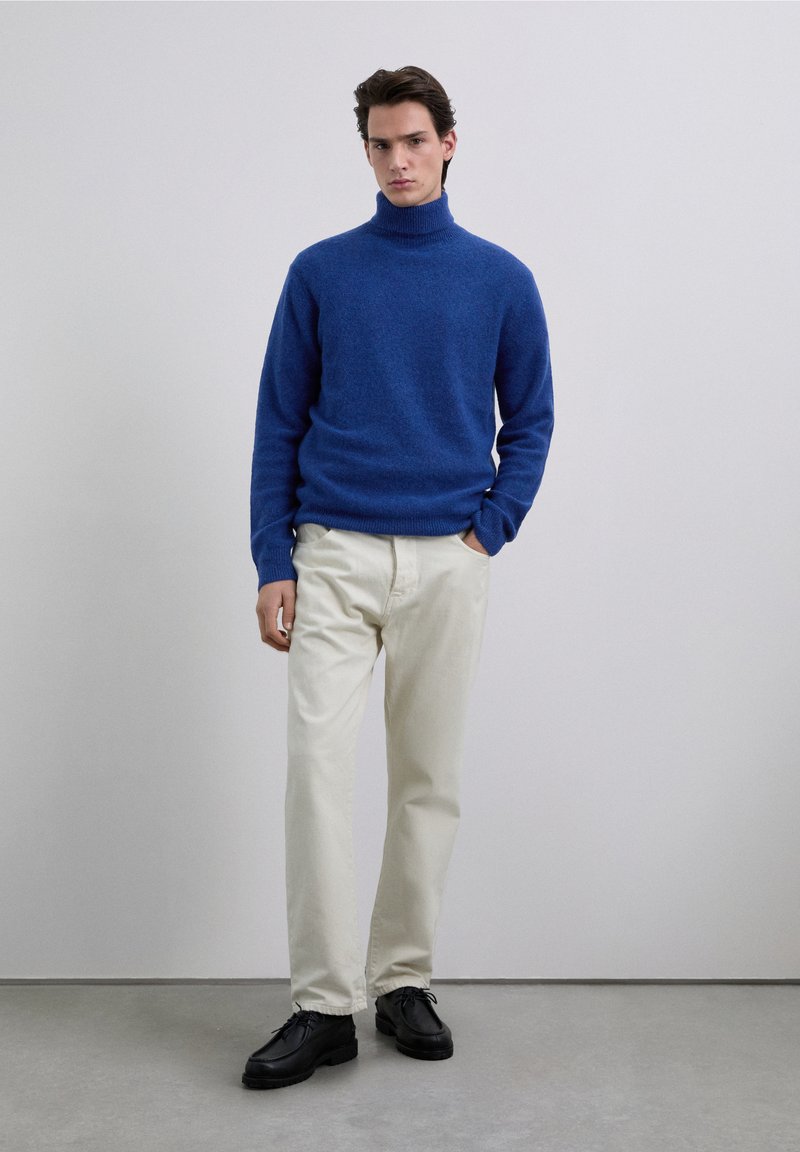 Maglione a collo alto blu, tessuto morbido, abbinato a pantaloni dritti color crema e scarpe nere stringate su sfondo neutro.