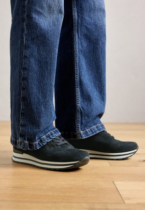 Isik kannab laiu siniseid teksaseid ja musti tossusid valgete tallaga, seistes puidust põranda peal.