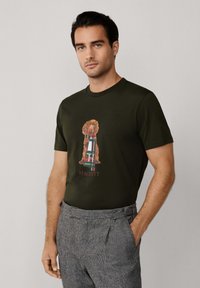 Camiseta de cuello redondo verde oliva con un diseño impreso de un perro con una bufanda a cuadros y el texto "HACKETT" debajo. Tejido de algodón suave.
