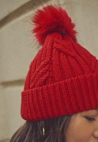 Bonnet en laine rouge tricoté avec un motif de tresses en relief et un pompon doux et duveteux sur le dessus. Poignet côtelé et surface texturée.