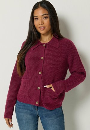 Femme aux longs cheveux foncés portant un cardigan bordeaux texturé boutonné avec des poches et un jean bleu, debout contre un mur uni.