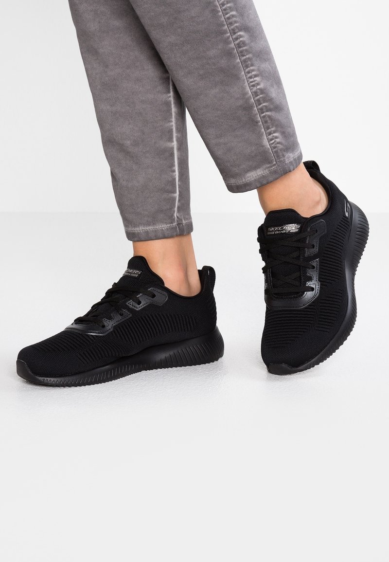 Personne portant des baskets Skechers BOBS noires avec une texture tricotée et des lacets noirs, se tenant sur un sol blanc avec un pantalon gris.