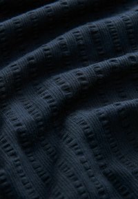 Granatowy materiał tekstylny z teksturowanym wzorem, z wypukłymi liniami i subtelnymi bruzdami, tworzącymi falisty wygląd. Miękka tkanina o gładkim wykończeniu.