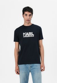 Svart bomulls T-skjorte med hvit "KARL LAGERFELD" logo foran, korte ermer og rund hals. Løs passform.