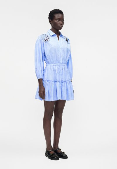 Γυναίκα που φοράει μπλε ριγέ φόρεμα με κοψίματα δαντέλας στους ώμους, μακριά μανίκια, φούστα με βολάν και μαύρες μπαλαρίνες Mary Jane, στέκεται ακίνητη.