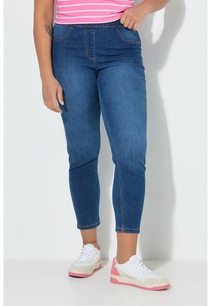 Frau trägt blaue Skinny-Jeans und weiße Sneaker mit pinken Akzenten, steht vor einer weißen Wand, die Hand auf der Hüfte ruhend.