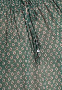 Tissu à motif floral vert avec des accents bleus et crème ; comprend un col froncé, des cordons de serrage et des boutons décoratifs sur le devant.