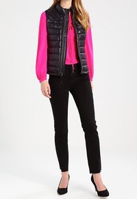 Veste matelassée noire sans manches avec un col montant portée sur un chemisier rose vif, associée à un pantalon noir et des talons noirs ; texture lisse et coupe ajustée.