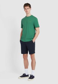 T-shirt verde a maniche corte con scollatura rotonda, abbinato a pantaloni corti blu navy, calzini bianchi e scarpe da ginnastica blu navy. Design semplice, texture liscia.