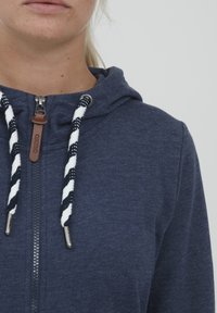 Mornarica zip pulover s kapuco, ki ima teksturirano tkanino, črtaste vrvi z usnjenimi detajli in standardno obliko kapuce.