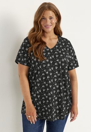 Lachende vrouw met lang rood haar draagt een zwarte blouse met korte mouwen met witte paardenbloempatronen en blauwe spijkerbroek tegen een effen achtergrond.