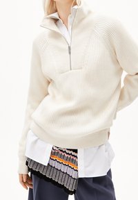 Pull-over en maille de couleur crème avec col demi-fermeture éclair, texture côtelée, et chemise superposée à motifs avec des rayures de différentes couleurs.