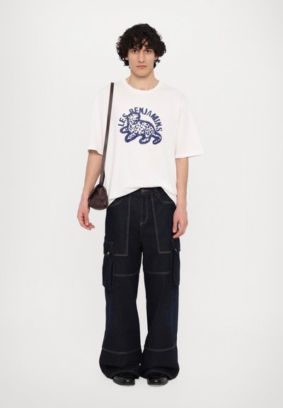 Λευκό T-shirt με μπλε γραφικά και κείμενο, συνδυασμένο με σκούρα τζιν cargo παντελόνια με τσέπες. Το μοντέλο κρατά μια μικρή καφέ τσάντα.