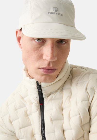 Beige quilted jacket with high collar and black zipper; logo on cap; textured fabric; close-up of face with light freckles.