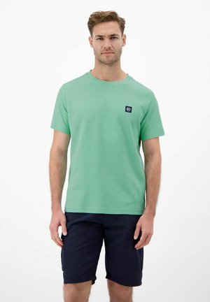 Mann trägt ein hellgrünes kurzärmeliges T-Shirt und dunkelblaue Cargoshorts, steht vor einem schlichten weißen Hintergrund.