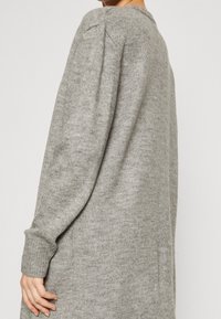 Šedý pletený svetr s dlouhými rukávy, kulatým výstřihem a žebrovanými manžetami. Má jemnou texturu a uvolněný střih.