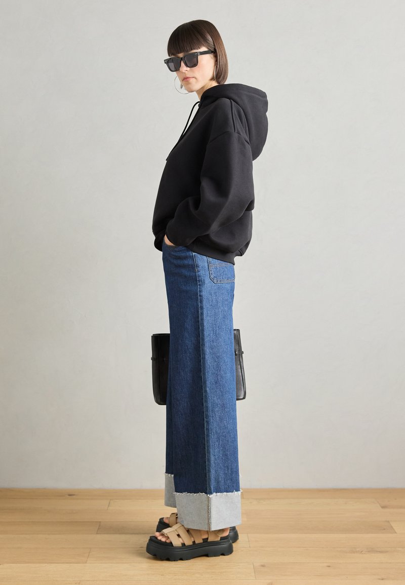 Schwarzer Kapuzenpullover, blaue weit geschnittene Jeans mit fransigem Saum, schwarze klobige Sandalen und übergroße Sonnenbrille. Eine schlichte schwarze Handtasche liegt an der Seite.