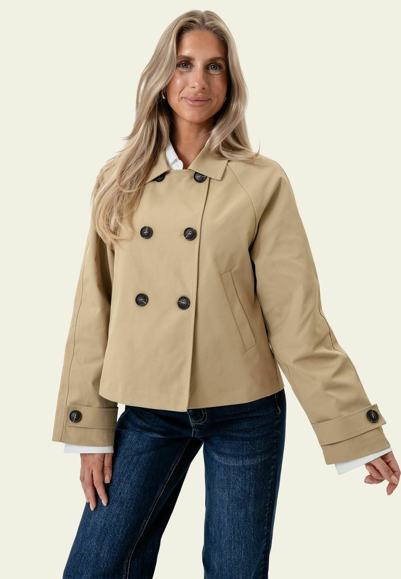 Femme aux longs cheveux blonds portant une veste beige à double boutonnage avec des boutons noirs et un jean bleu foncé, debout devant un fond clair.