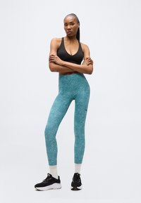 Γυναίκα όρθια με σταυρωμένα τα χέρια, φοράει μαύρο αθλητικό σουτιέν, τιρκουάζ υφασμάτινο κολάν, λευκές κάλτσες και μαύρα αθλητικά παπούτσια σε λευκό φόντο.