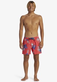 Red swim shorts with blue floral patterns, elastic waistband with drawstring, and side pockets. Model shown is standing barefoot.