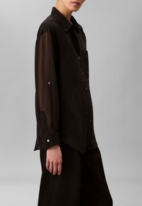 Sort skjorte med knapper, der har en ensfarvet krop og gennemsigtige lange ærmer med en enkelt brystlomme og metal knappedetaljer. Glat tekstur.