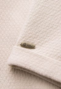 Tissu texturé crème avec une petite étiquette en métal portant la mention Ombre Heritage attachée près d’un bord plié.