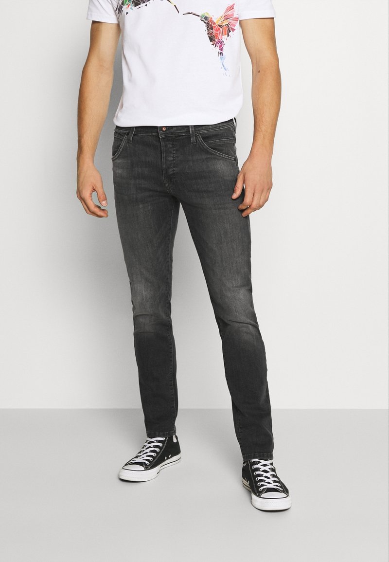 Jean en denim noir, coupe slim, avec un léger effet délavé et des plis d'usure. Style avec un t-shirt blanc à motif et des baskets noires.