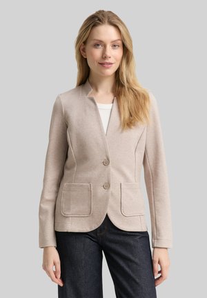 Kvinde med langt blond hår iført beige knappet blazer over hvid top og mørke jeans, stående mod grå baggrund.