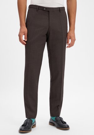 Mann trägt dunkelbraune Anzughose, türkise Socken und schwarze Tassel-Loafer, steht vor einem schlichten Hintergrund.
