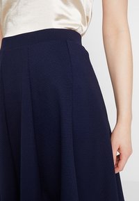 Mörkblå texturerad kjol med hög midja, med veck och ett slätt, glänsande tyg upptill. Hand placerad på midjan.