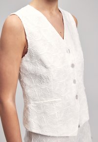 White embroidered vest featuring a V-neck, five buttons, and two front pockets. The texture has a floral pattern with leaf-like designs.
