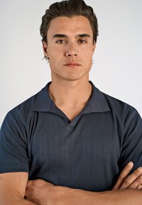 Nuori mies, jolla on tumma hius, seisoo kädet ristissä ja katsoo suoraan eteenpäin yksinkertaista taustaa vasten, päällä tummansininen tekstuuroitu polo-paita.