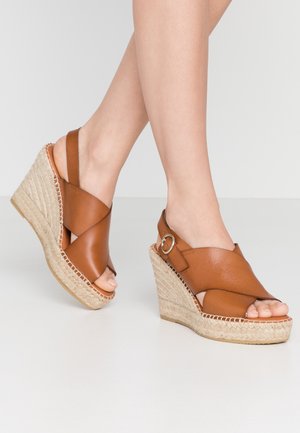Pies con sandalias de cuña de cuero tostado con tiras cruzadas y suelas plataforma de cuerda de yute sobre una superficie blanca.