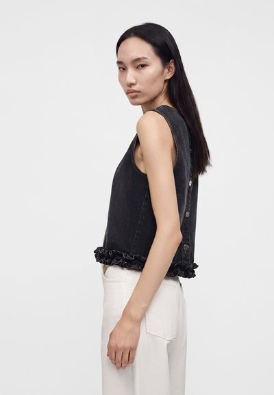 Femme aux longs cheveux foncés portant un top en denim noir sans manches avec des boutons dans le dos et un ourlet à volants, associé à un pantalon blanc, sur fond uni.