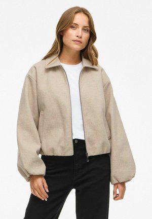 Beige cropped jack in zachte stof, met een ritssluiting aan de voorkant, oversized mouwen en zijzakken. Draagt over een wit shirt en zwarte spijkerbroek.
