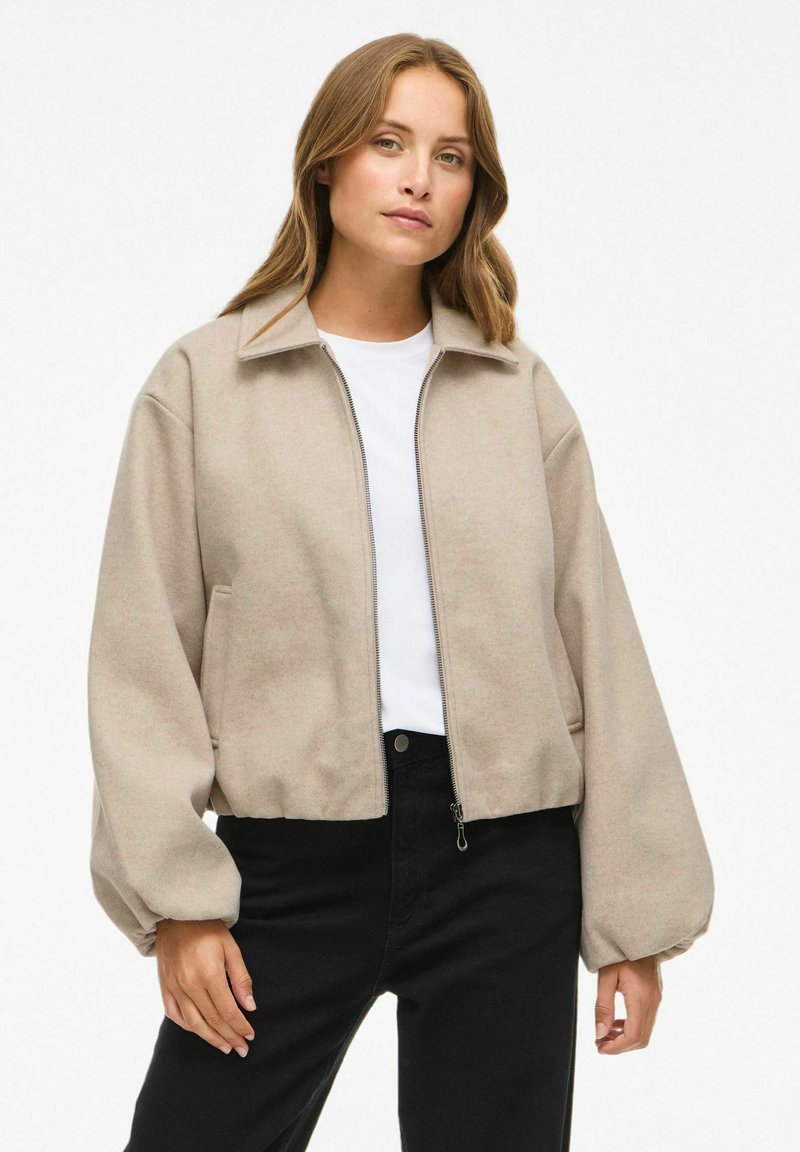 Beidzis krēmkrāsas apgrieztais žakets mīkstā audumā, ar priekšējo rāvējslēdzēju, pārmērīgiem piedurknēm un sānu kabatām. Uzvilkts virs baltas krekla un melnām džinsu biksēm.