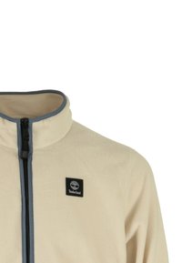 Giacca in pile beige con colletto alto, zip frontale, rifiniture blu e un'etichetta del logo Timberland nera sul lato sinistro del petto. Tessuto morbido.