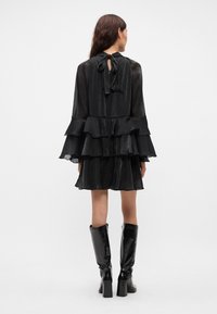 Must mustriga kleit, millel on läbipaistev kangas, pikad varrukad, volangide disain ja sidumisega kaelus. Komplekteeritud läikivate, põlvekõrguste mustade saapastega.