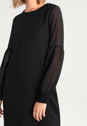 Frau trägt ein schwarzes Kleid mit transparenten langen Ärmeln und elastischen Bündchen, dargestellt von der Schulter bis zur Mitte des Oberschenkels vor einfarbigem Hintergrund.