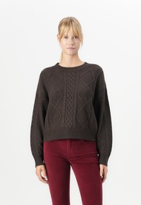 Ženska z blond lasmi nosi temno rjav pulover z pleteno obleko in hlače iz burgundske kordine z visokim pasom, stoji ob belem ozadju.