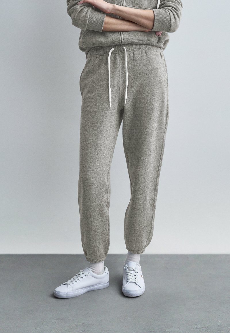 Άτομο που φοράει ανοιχτό γκρι φόρμα, λευκές κάλτ σες και λευκά αθλητικά παπούτσια, στέκεται με τα χέρια σταυρωμένα πάνω σε απλό γκρι φόντο.