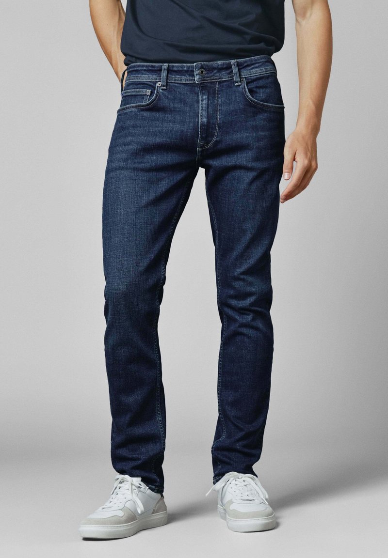 Tamsiai mėlyni džinsai su tiesiu kirpimu, penkių kišenių dizainu ir subtiliomis siuvimo detalėmis. Dėvimi su baltais sportbačiais.