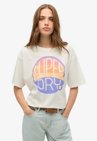 Camiseta de manga corta color crema claro con un gran logo circular multicolor "SUPER DRY" en naranja, rosa y púrpura. Corte casual con cuello redondo.