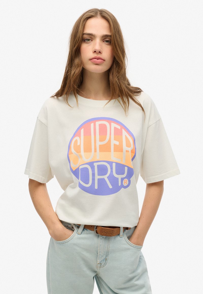 Camiseta de manga corta color crema claro con un gran logo circular multicolor "SUPER DRY" en naranja, rosa y púrpura. Corte casual con cuello redondo.