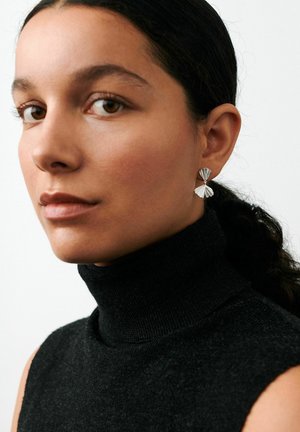 Femme aux cheveux foncés ramenés en arrière, portant un col roulé noir sans manches et des boucles d'oreilles pendantes argentées en forme d'éventail, légèrement tournée vers la gauche.