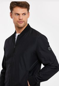 Schwarze Bomberjacke mit durchgehendem Reißverschluss, gerippten Bündchen und Kragen, zwei seitlichen Taschen und einem runden Logo-Patch am linken Ärmel.