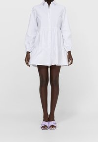 Femme portant une longue robe chemise blanche à boutons avec des manches bouffantes et des mules ouvertes à talons légers violets, se tenant devant un fond uni.